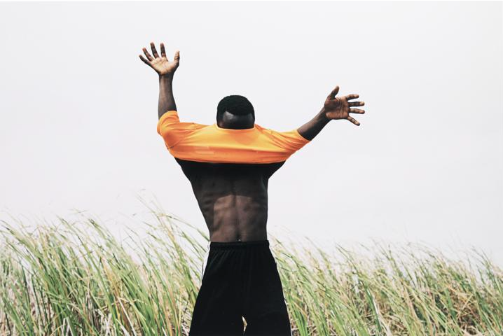 Man in a field of grass with his hands splayed and shirt around his neck
