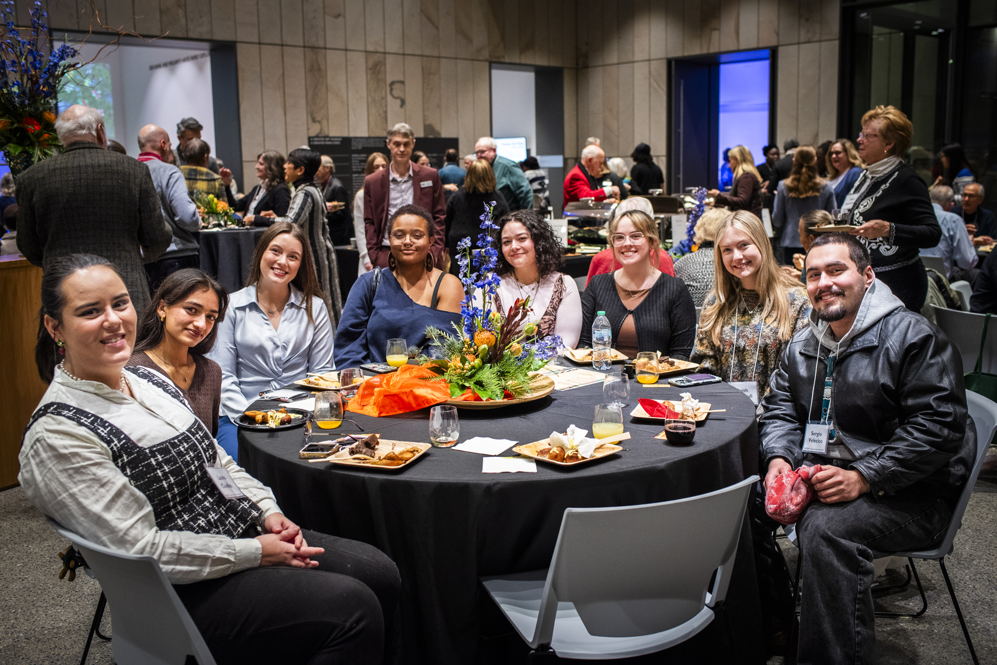 A group of young people sitting around a table with a black tablecloth at an event reception in a lobby with sandstone walls.